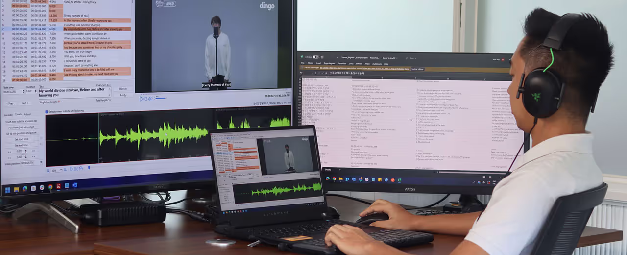 A man wearing headphones works at a desk with multiple monitors displaying subtitling and translation software. He is editing subtitles for a video of a singer performing on a stage, with waveforms and text synchronization tools visible on the screens.