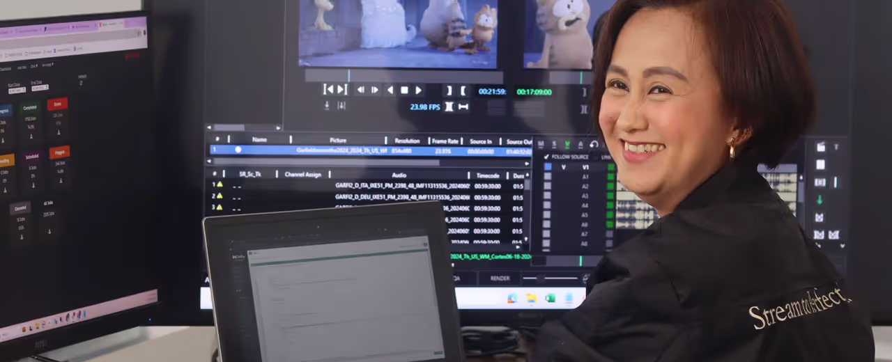 A woman wearing a jacket works at a desk with multiple monitors displaying video editing and quality assurance software. The screens show a scene from an animated film, audio tracks, and workflow progress indicators.